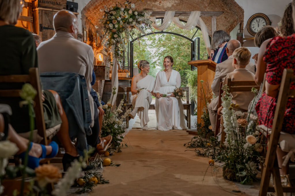 Bruiden zitten samen vooraan de ceremonie en gasten aan het gangpad zijn zichtbaar