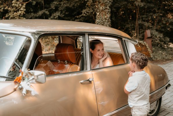 Bruid achter in de bruidsauto met bruidsjonker die door het raampje kijkt